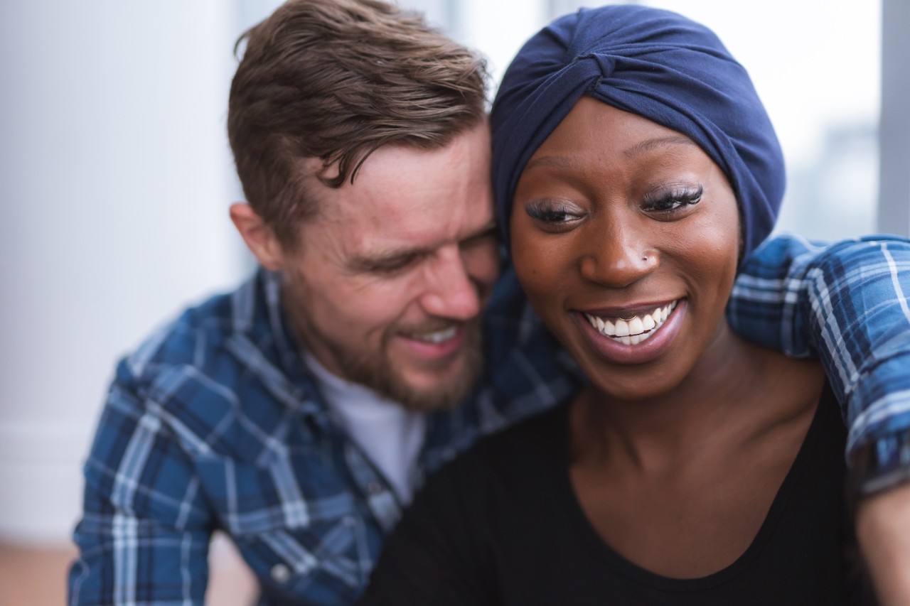 A handsome caucasian man is hugging his stunning African American wife who is recovering from cancer. They are smiling and feeling optimistic.